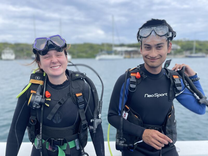 Two people in scuba gear are posing for a photo. They are on a boat, with water and land visible in the background. Both divers are wearing black wetsuits and scuba equipment, including masks and tanks. The person on the left is smiling broadly, while the person on the right has a more subtle smile. The sky is overcast.